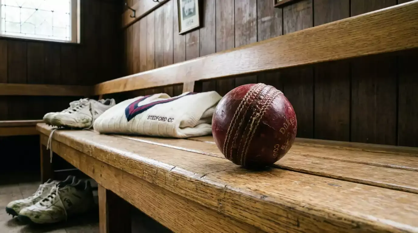 Worn red cricket ball resting on the wooden bench of an empty pavilion at a quiet county ground